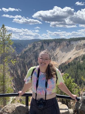 A girl with a tie-dye crop top and black leggings stands facing the camera with her back to Yellowstone's Grand Canyon